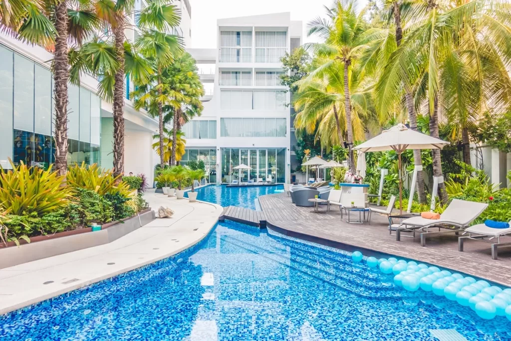 Imagem de um resort com piscina de água azul, cercada por palmeiras e áreas de lazer com espreguiçadeiras. O hotel tem uma fachada moderna, com janelas grandes e mesas ao ar livre perto da piscina.