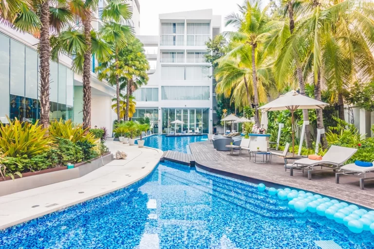 Imagem de um resort com piscina de água azul, cercada por palmeiras e áreas de lazer com espreguiçadeiras. O hotel tem uma fachada moderna, com janelas grandes e mesas ao ar livre perto da piscina.