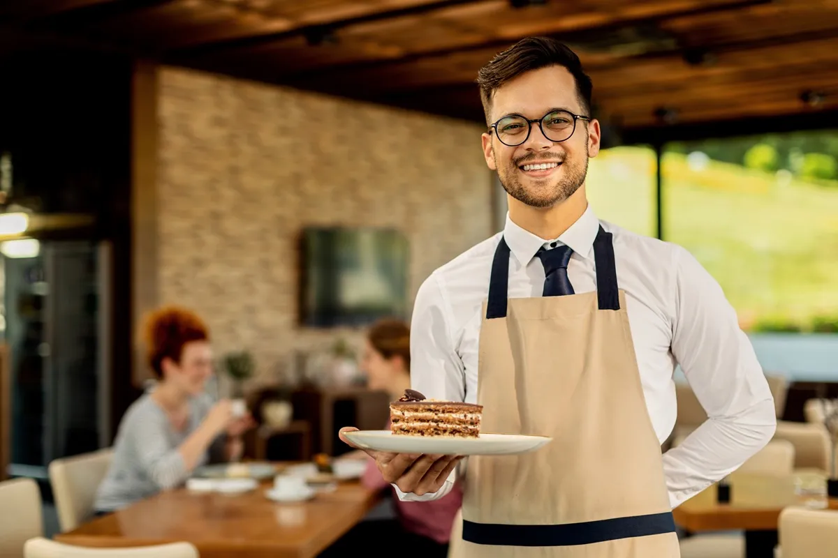 Garçom sorrindo com óculos e avental, segurando um prato com uma fatia de bolo em restaurante moderno, com ambiente acolhedor e outras pessoas ao fundo.