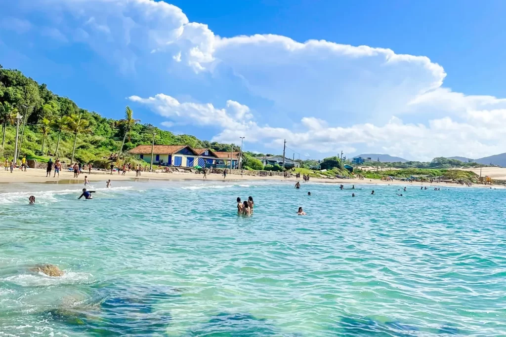 Praia tropical brasileira com mar cristalino, casas coloridas e banhistas aproveitando o dia ensolarado.