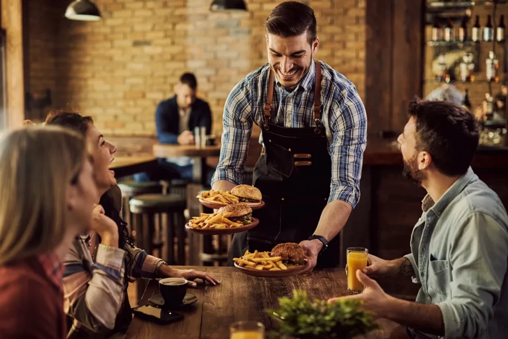 Garçom sorrindo com camisa xadrez e avental preto, servindo dois pratos com hambúrgueres e batatas fritas para um casal em um restaurante de ambiente rústico e moderno, com iluminação suave.