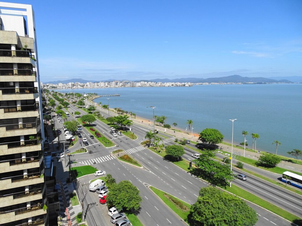 Vista aérea da Avenida Beira-Mar Norte em Florianópolis com mar calmo, prédios urbanos, pista arborizada e céu azul