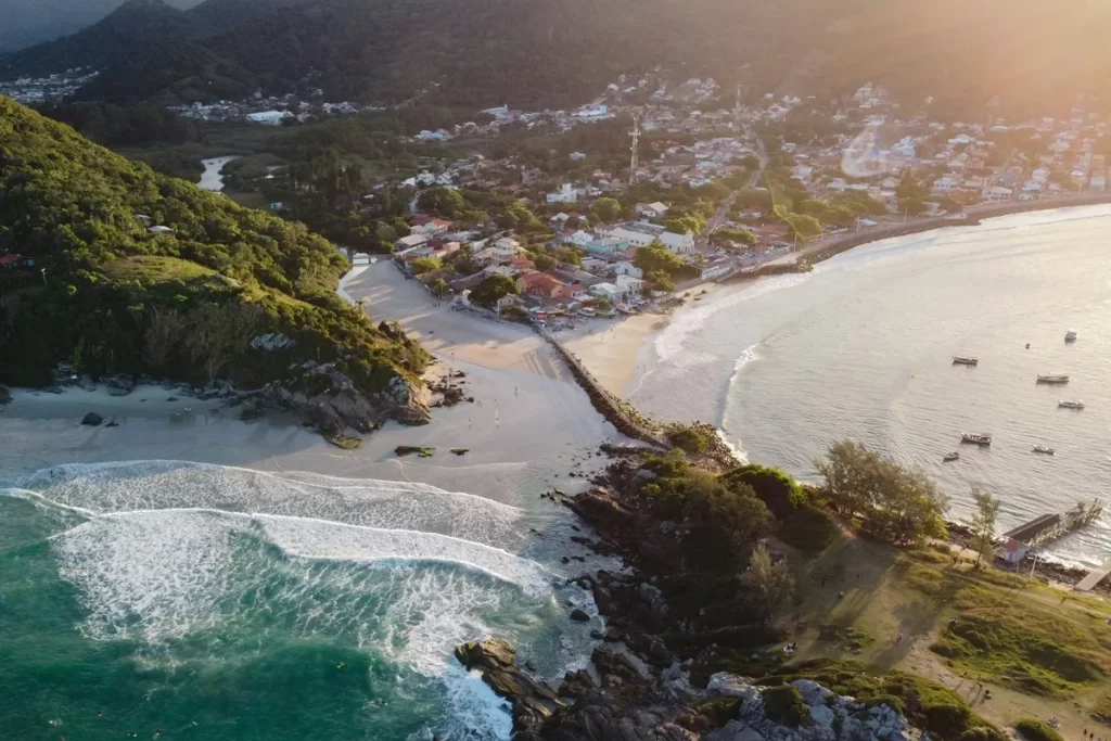Imagem aérea de vila costeira com duas praias, mar calmo com barcos, vegetação densa e morros ao fundo ao entardecer.