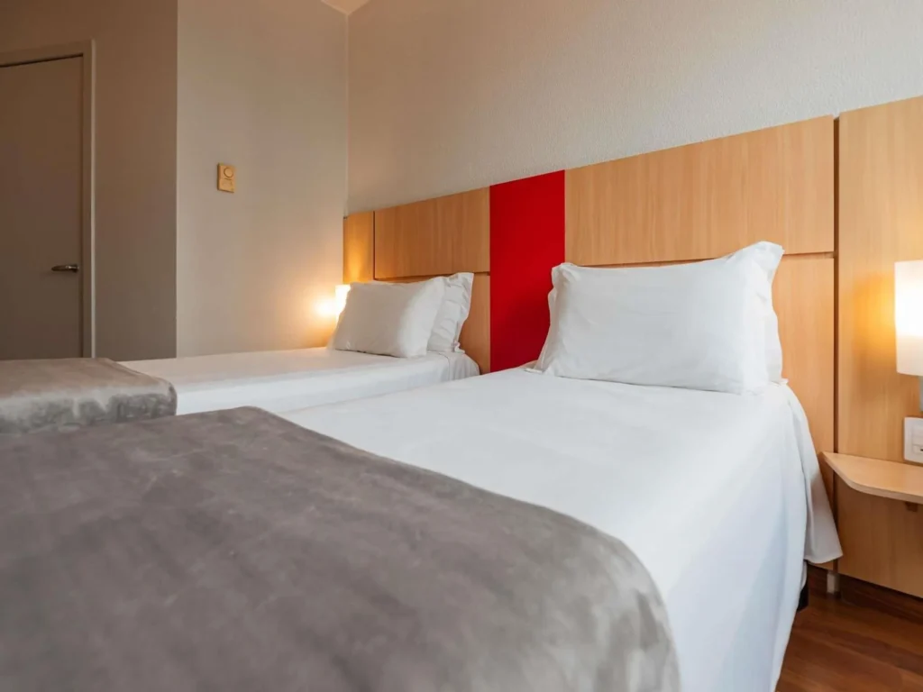 Quarto de hotel em Florianópolis com duas camas de solteiro, roupa de cama branca, iluminação aconchegante e decoração moderna