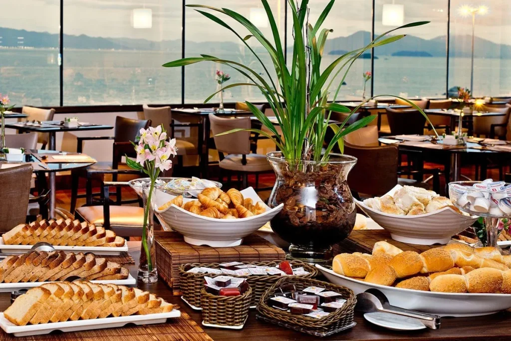 Buffet de café da manhã em hotel de Florianópolis com pães, croissants, frutas e vista panorâmica para o mar
