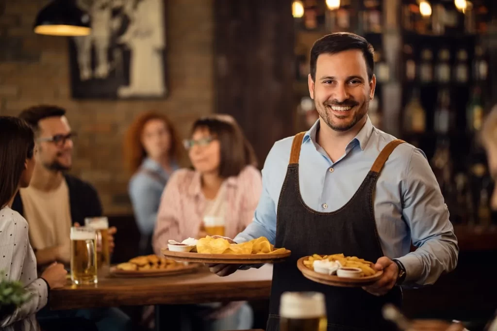 Garçom sorrindo, vestindo camisa azul e avental preto, segurando dois pratos com petiscos e molhos em bar com ambiente aconchegante e iluminado, com clientes ao fundo.