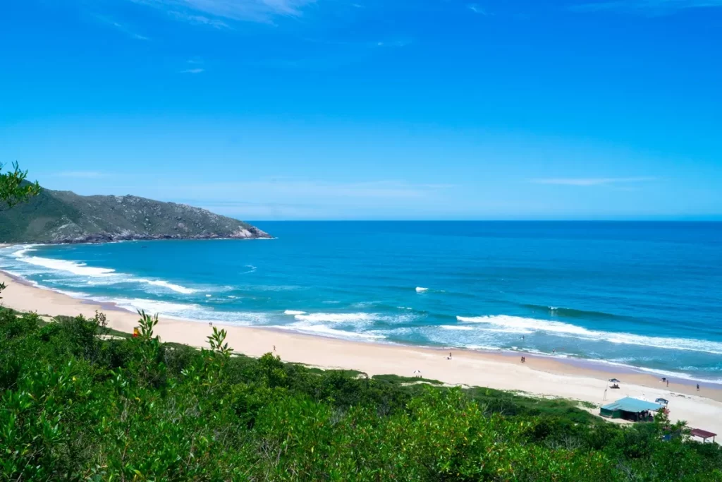 Vista panorâmica de uma praia tranquila com águas azul-turquesa, areia clara e vegetação ao redor, oferecendo uma paisagem relaxante e natural.