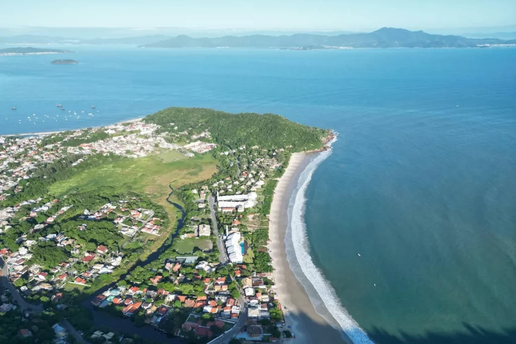 Vista aérea mostrando a praia de areia branca, o mar azul e a área urbana próxima, com casas, ruas e vegetação ao redor.