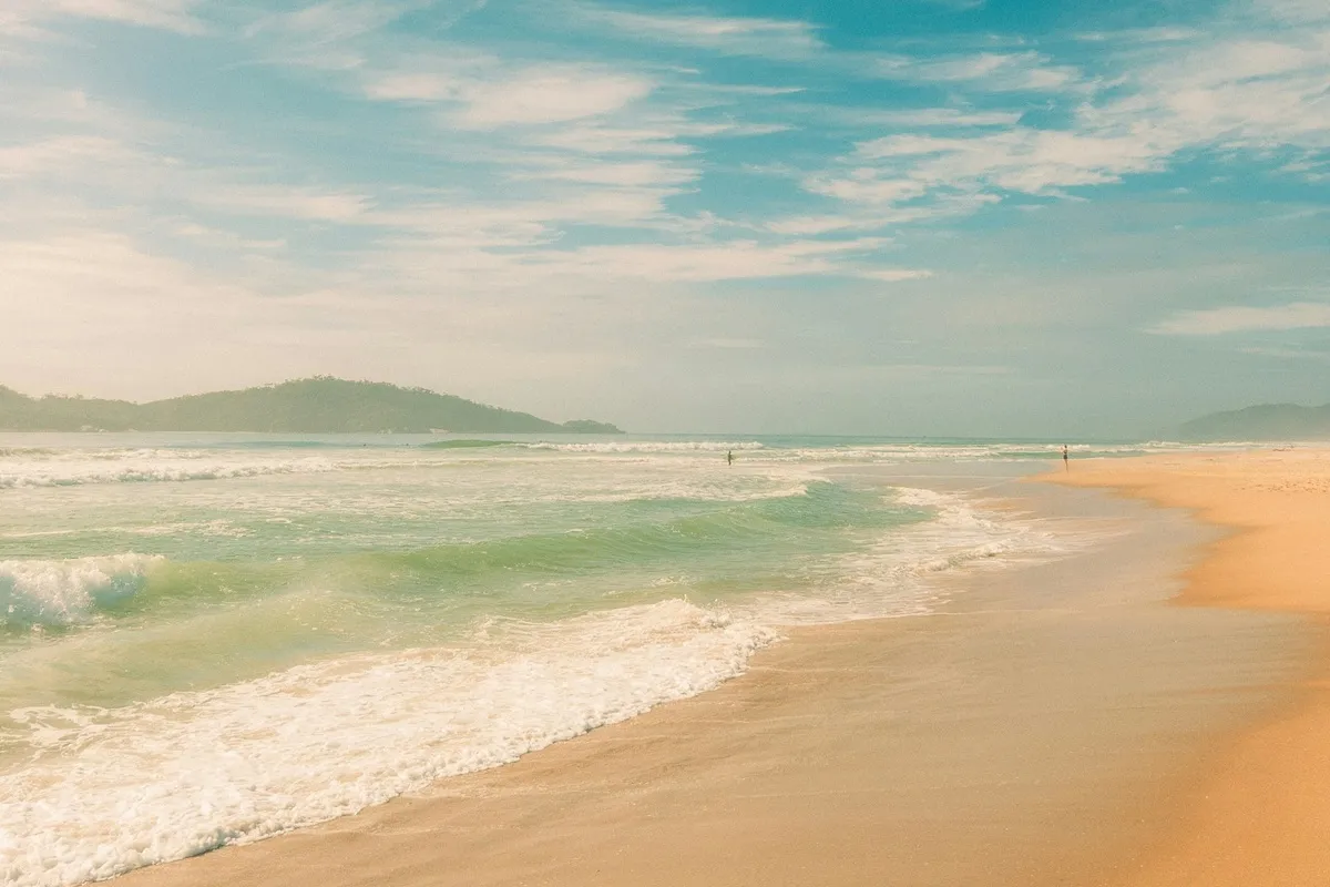 Vista tranquila de uma praia com águas verde-azuladas, ondas suaves e céu azul claro, com uma pessoa caminhando ao longo da costa