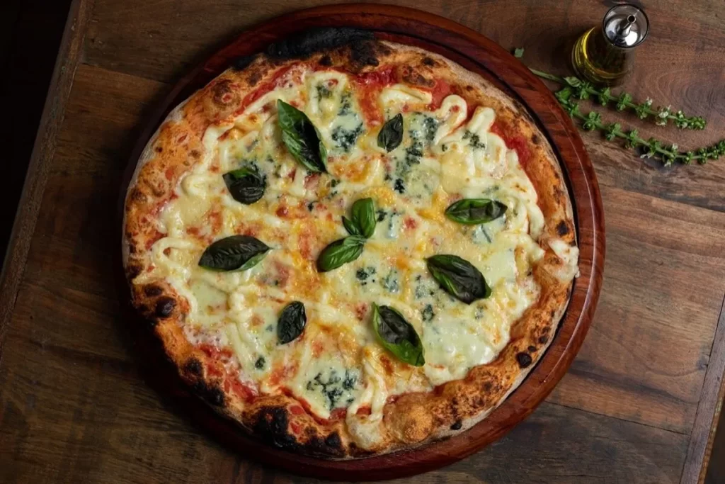 Pizza artesanal de quatro queijos com borda levemente tostada, molho de tomate, folhas frescas de manjericão e servida em tábua de madeira sobre mesa rústica.