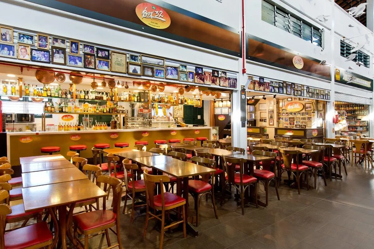 Interior do restaurante Box 32 no Mercado Público de Florianópolis, com balcão iluminado, diversas garrafas expostas, bancos altos vermelhos e várias mesas de madeira com cadeiras estofadas.