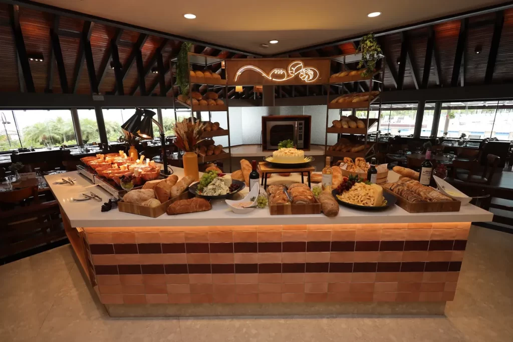 Mesa de buffet com pães artesanais, queijos, frutas e vinhos em ambiente elegante de restaurante.