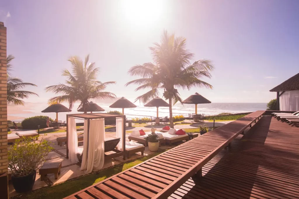 Espaço de lazer à beira-mar com espreguiçadeiras, gazebos, palmeiras e vista para o oceano ao pôr do sol.