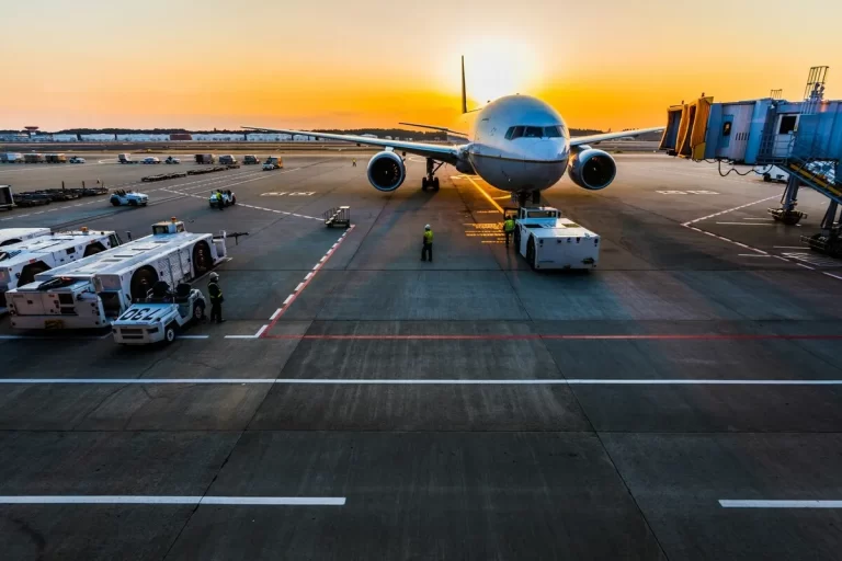 Avião no pátio do aeroporto ao pôr do sol, com equipamentos de solo e finger de embarque, simbolizando viagens aéreas e a proximidade de hotéis.