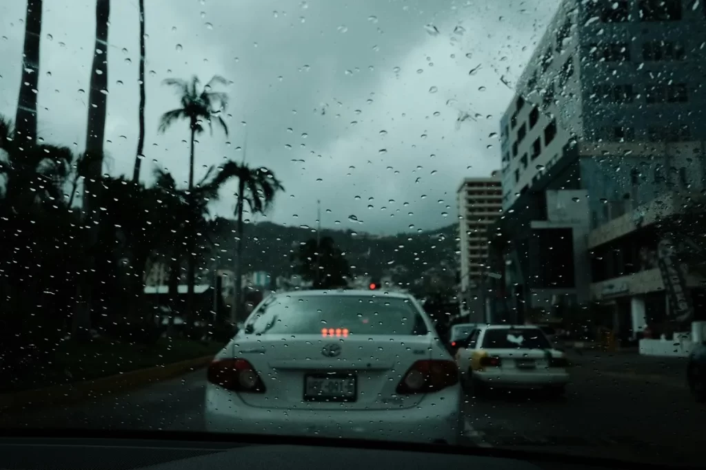 Vista de um carro no trânsito em Florianópolis, com gotas de chuva no para-brisa, prédios e árvores ao fundo em dia nublado