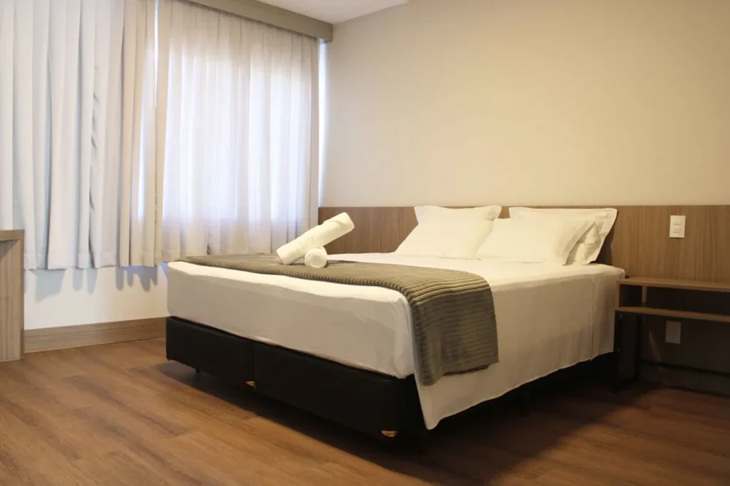 Quarto de hotel no centro de Florianópolis com cama de casal, piso de madeira, cortinas claras e decoração moderna.
