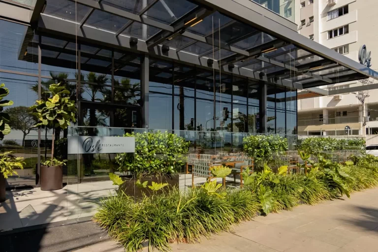 Fachada envidraçada do Oslé Restaurante com teto em estrutura metálica, plantas tropicais em vasos e área de mesas ao ar livre