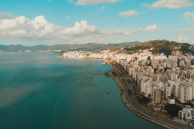Vista panorâmica da cidade de Florianópolis com o litoral à esquerda, prédios ao longo da costa e montanhas ao fundo, sob céu azul com algumas nuvens