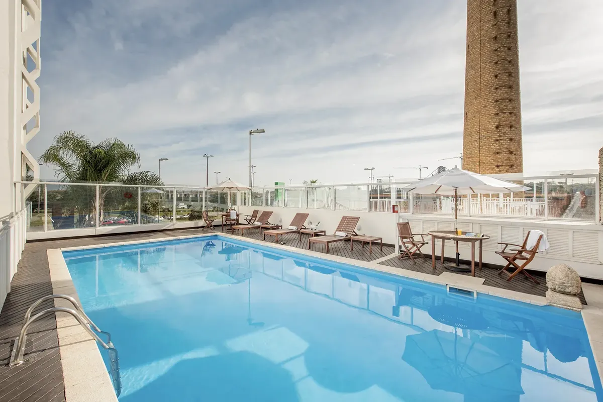 Piscina externa em terraço com espreguiçadeiras de madeira, guarda-sóis brancos e vista urbana ao fundo