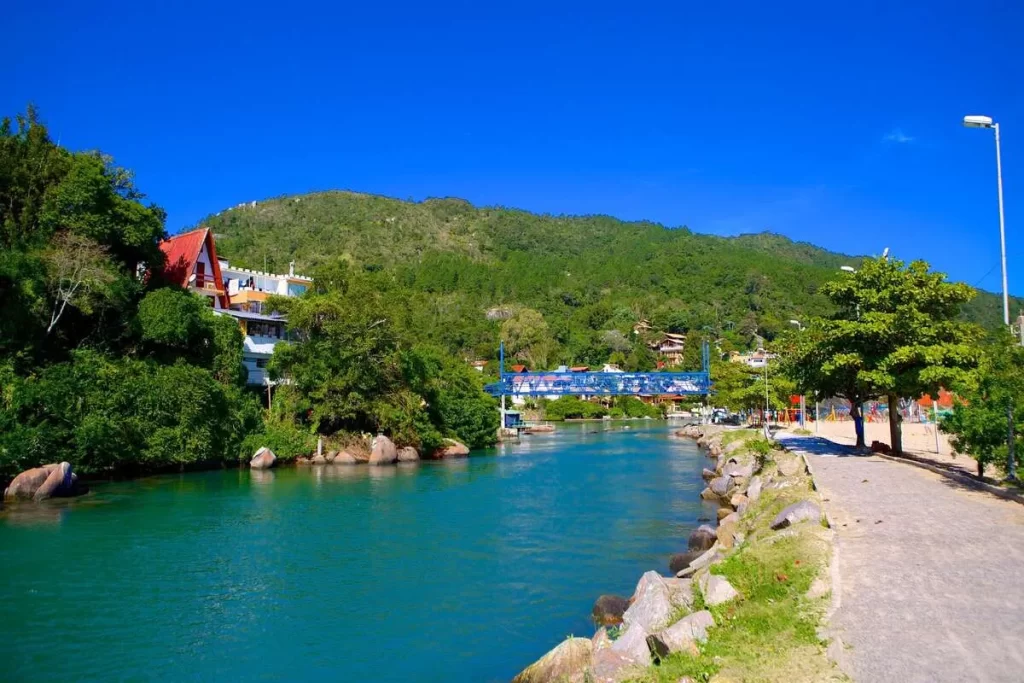 Canal de águas azuis-turquesa conectando a Lagoa da Conceição ao oceano na Barra da Lagoa, Florianópolis, com ponte azul, vegetação exuberante e céu azul intenso.