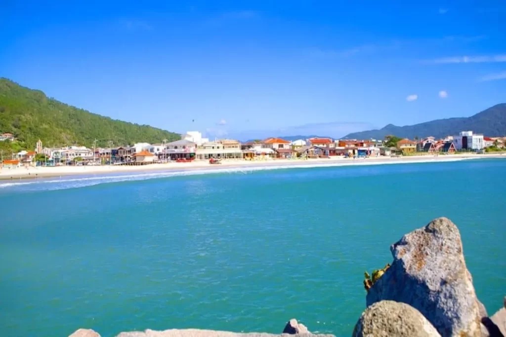 Vista panorâmica da praia de Barra da Lagoa, Florianópolis, com mar azul-turquesa, areia branca, construções costeiras e morros verdes sob céu ensolarado.