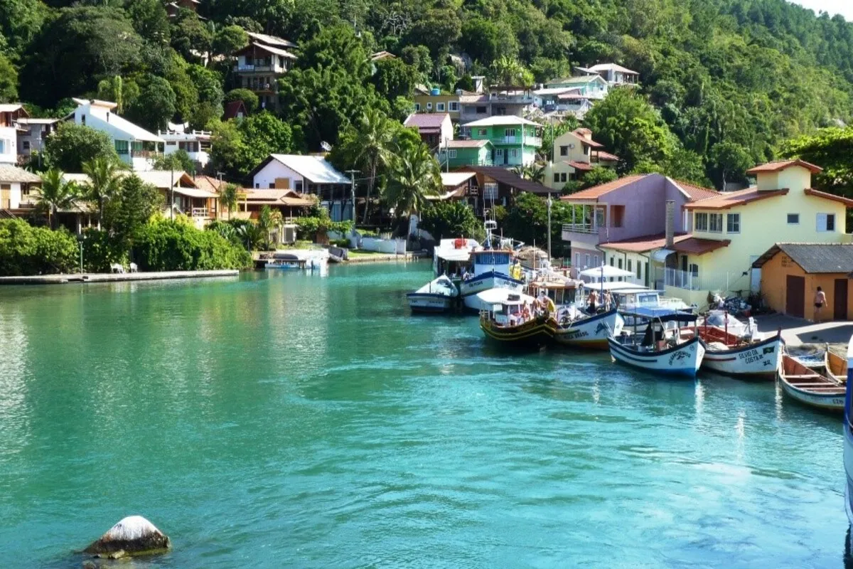 Barcos de pesca coloridos atracados em águas verdes-turquesa, com casas na encosta de morros verdes na Barra da Lagoa, Florianópolis.