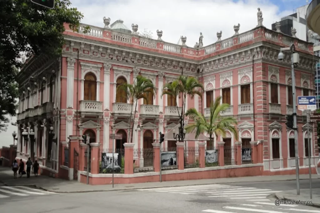 Edifício histórico rosa com detalhes ornamentados no Centro Histórico de Florianópolis, palmeiras e rua com pedestres.