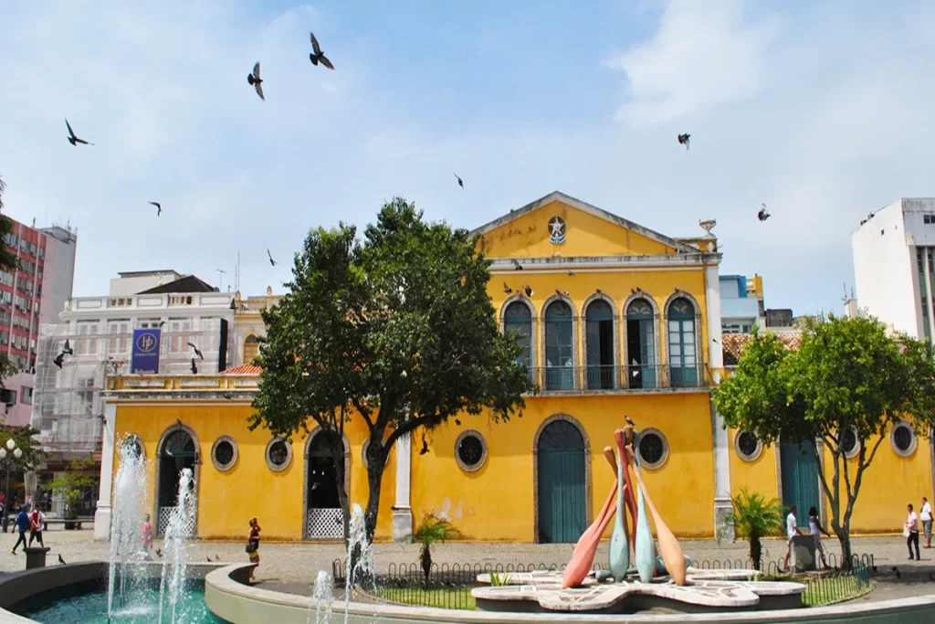 Edifício amarelo do Centro Histórico de Florianópolis com fonte e esculturas, pombos voando, céu azul.