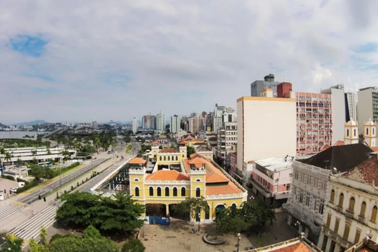 Edifício amarelo do Centro Histórico de Florianópolis com fonte e esculturas, pombos voando, céu azul.