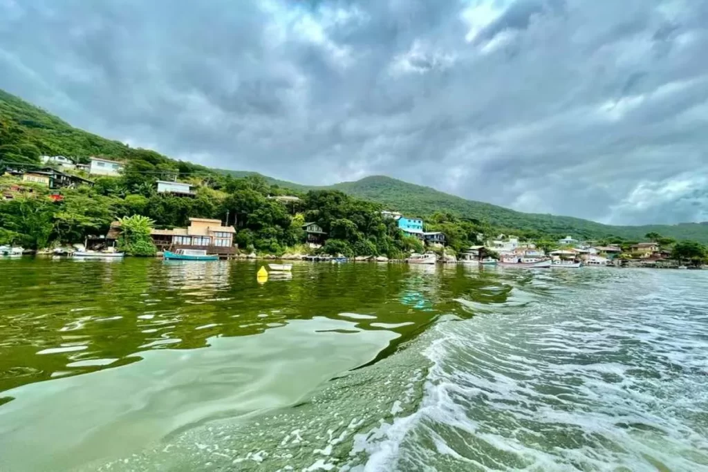 Casas coloridas e barcos na margem da Lagoa da Conceição em Costa da Lagoa, Florianópolis, sob céu nublado e vegetação exuberante.