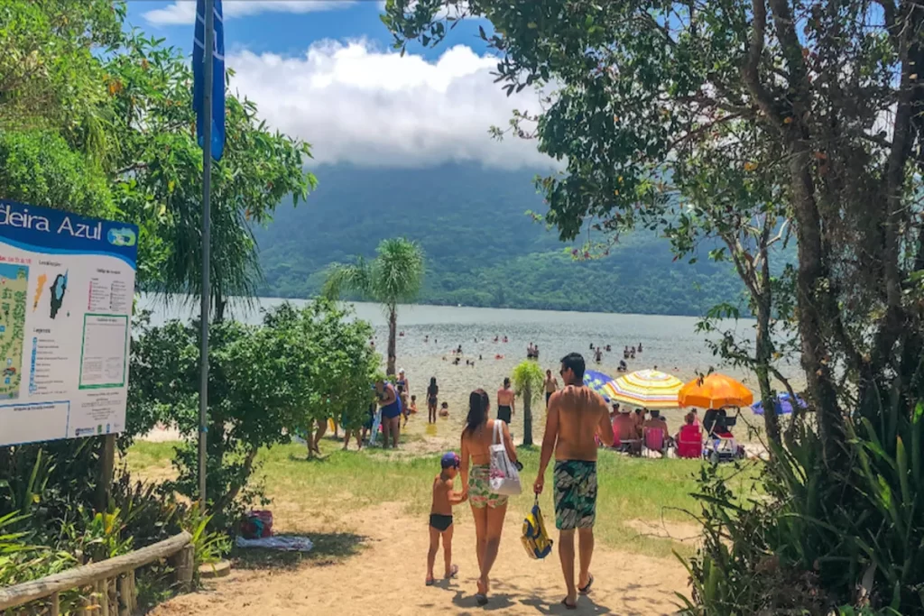 Família caminhando na areia em direção à Lagoa do Peri, com pessoas na água e montanhas ao fundo. Praia e natureza em Florianópolis.