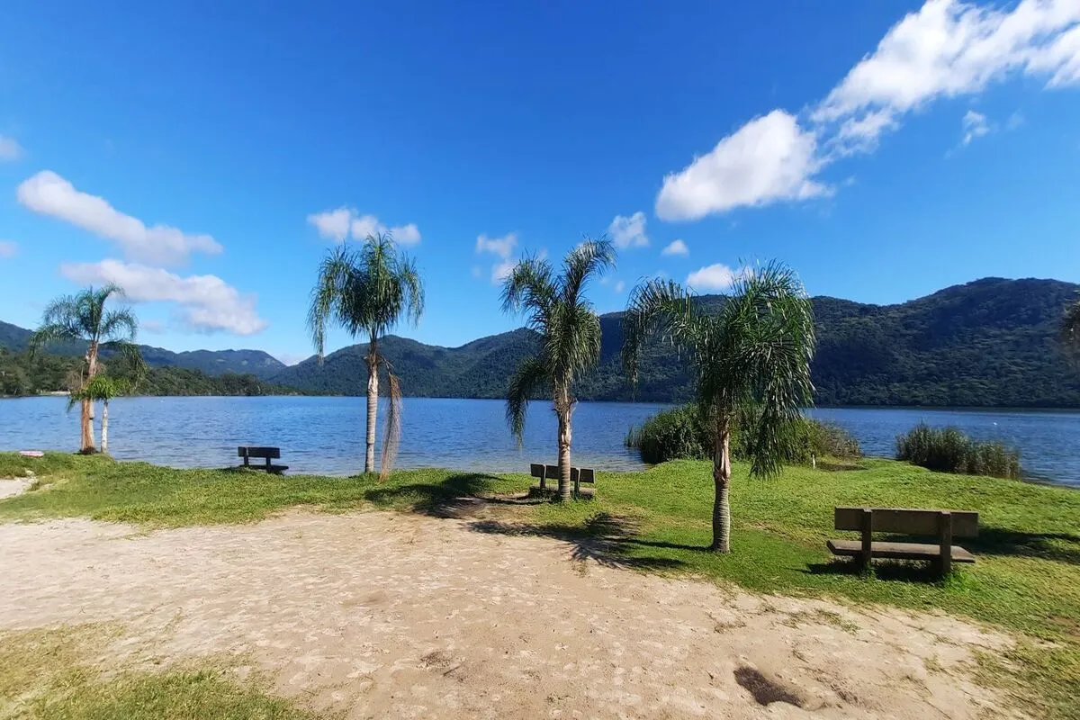 Paisagem panorâmica da Lagoa do Peri em dia ensolarado, com palmeiras, bancos rústicos e montanhas ao fundo. Natureza e tranquilidade.