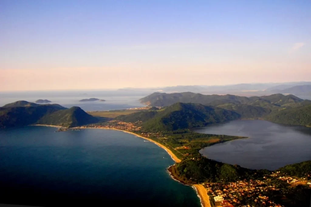 Vista aérea de uma grande lagoa de água escura, separada do oceano por uma faixa de areia e vegetação em uma área montanhosa.
