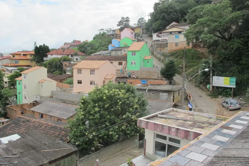Vista de um bairro residencial no Morro da Cruz, com casas coloridas e telhados variados subindo uma encosta arborizada, uma rua estreita e vegetação exuberante típica da região.