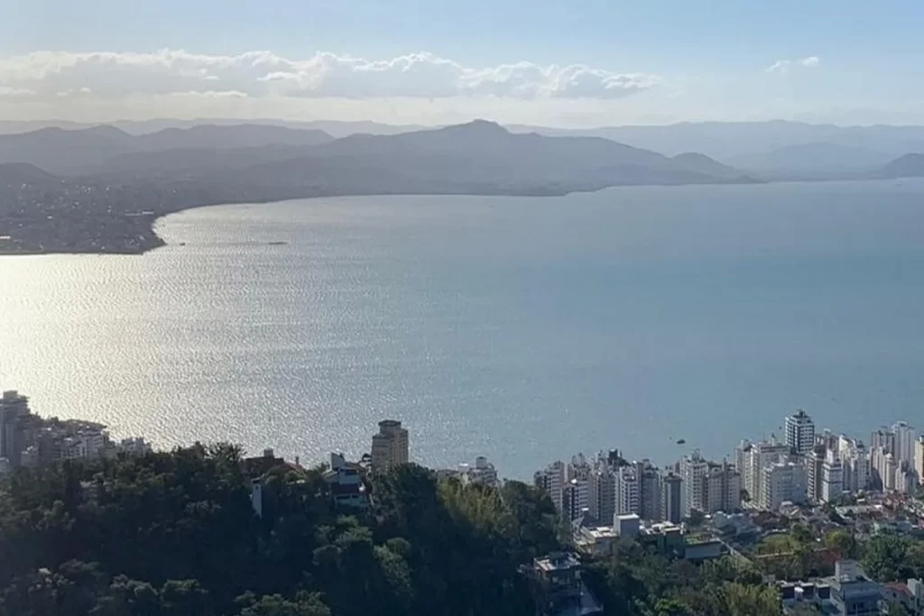 Vista aérea deslumbrante do Morro da Cruz em Florianópolis, mostrando a baía cintilante, a expansão urbana com seus edifícios e as montanhas ao fundo sob um céu claro.