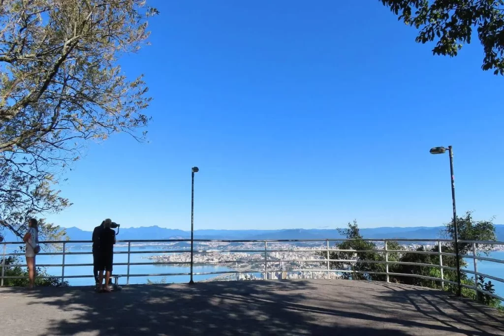 Duas pessoas no mirante do Morro da Cruz em Florianópolis, olhando para a vasta paisagem da baía, a cidade abaixo e as montanhas distantes, sob um céu azul claro.
