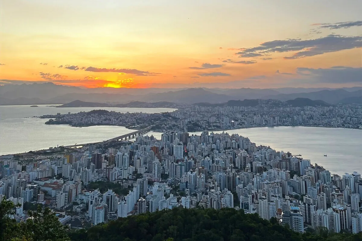 Vista panorâmica do Morro da Cruz ao pôr do sol, mostrando a cidade de Florianópolis iluminada, a Ponte Hercílio Luz, as baías cintilantes e montanhas distantes, tudo sob um céu com tons alaranjados e rosados.