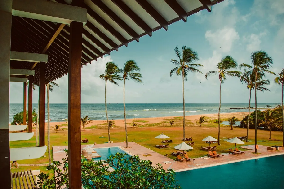 Um quarto de hotel moderno e elegante com uma enorme janela de vidro que oferece uma vista deslumbrante do oceano azul, uma praia movimentada com coqueiros e uma área urbana costeira, exemplificando o tipo de acomodação espaçosa e com vista que o Novotel Florianópolis pode oferecer.