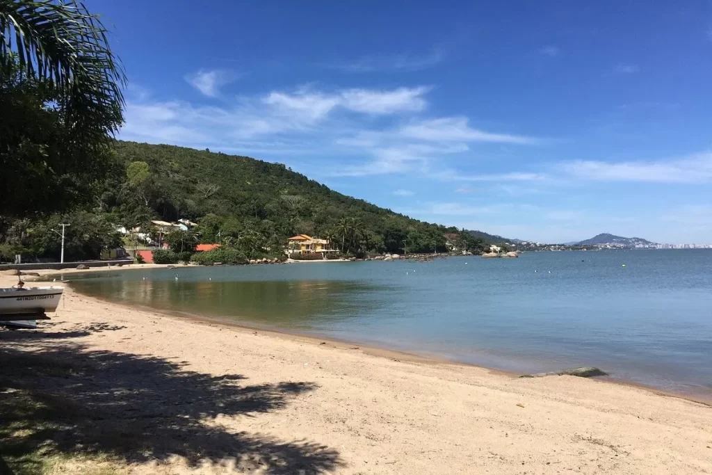 Uma vista panorâmica da Lagoa da Conceição, em Florianópolis, com uma praia de areia clara em primeiro plano, águas calmas, e uma colina coberta por vegetação densa. Ao fundo, uma cidade e uma montanha podem ser vistas sob um céu azul.