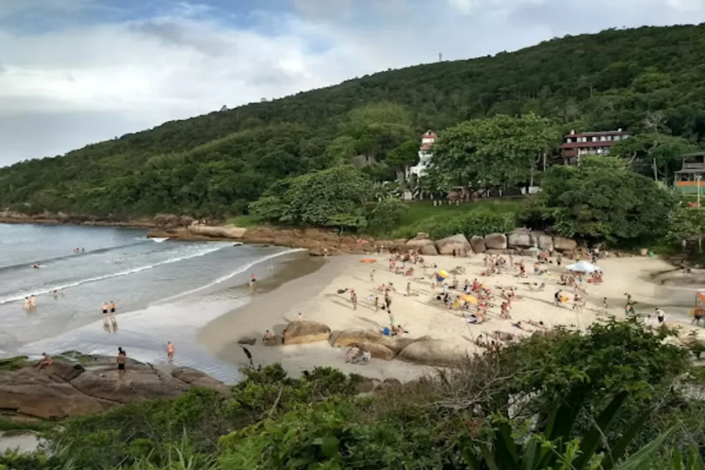 Uma vista panorâmica da Prainha da Barra da Lagoa, com uma praia de areia clara, rochas e mar com pequenas ondas. Uma colina verdejante coberta de mata atlântica e uma construção com um farol de mentira podem ser vistas ao fundo.
