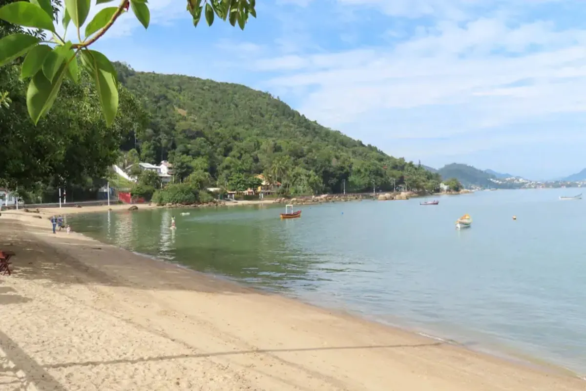 Uma vista da praia da Barra da Lagoa, com uma faixa de areia dourada em primeiro plano, águas calmas e pequenas embarcações. O fundo mostra uma colina densamente arborizada, casas e uma paisagem urbana distante.