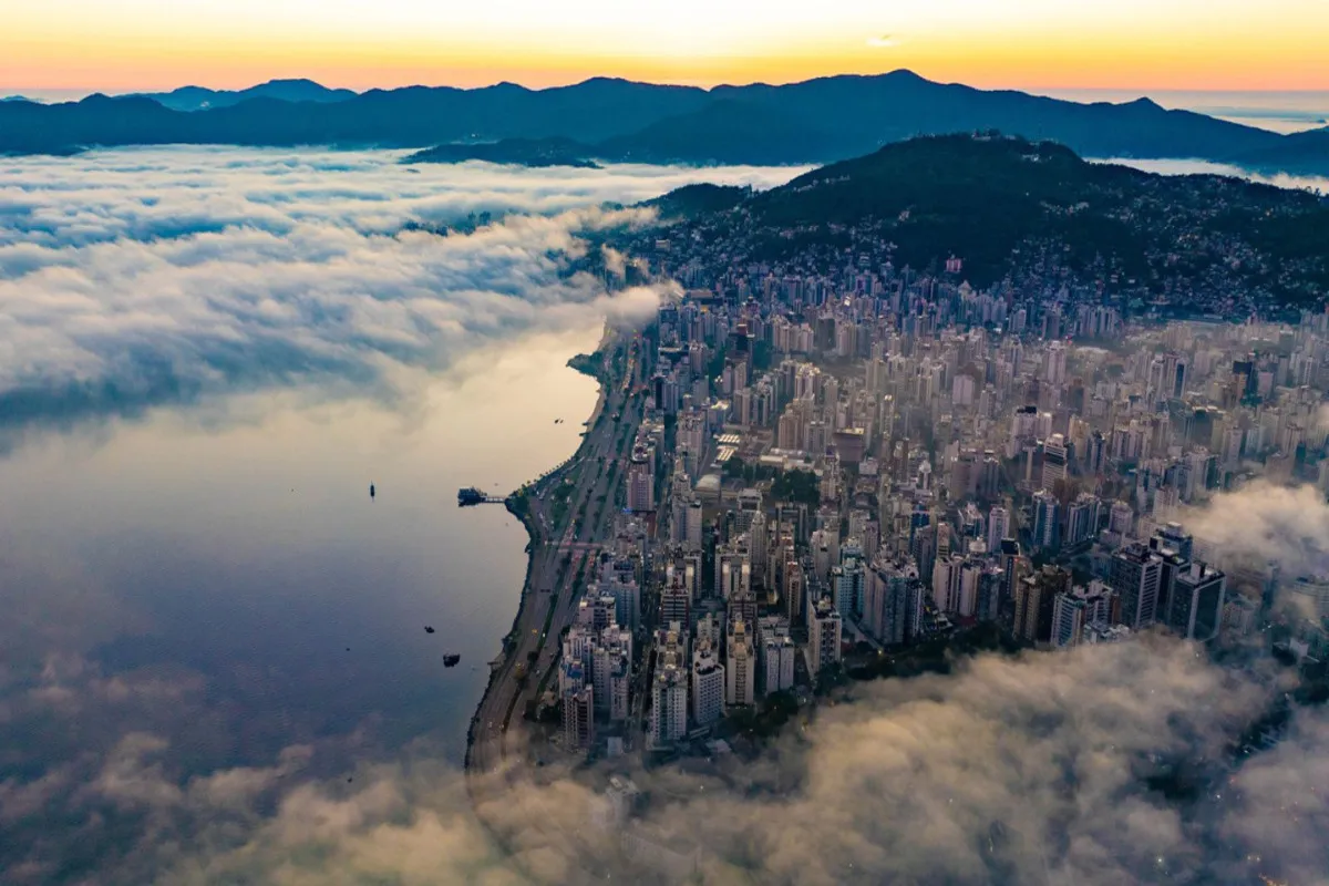 Uma vista aérea da cidade de Florianópolis, com a orla e edifícios parcialmente cobertos por uma densa camada de neblina que se estende sobre a baía e o mar ao pôr do sol.