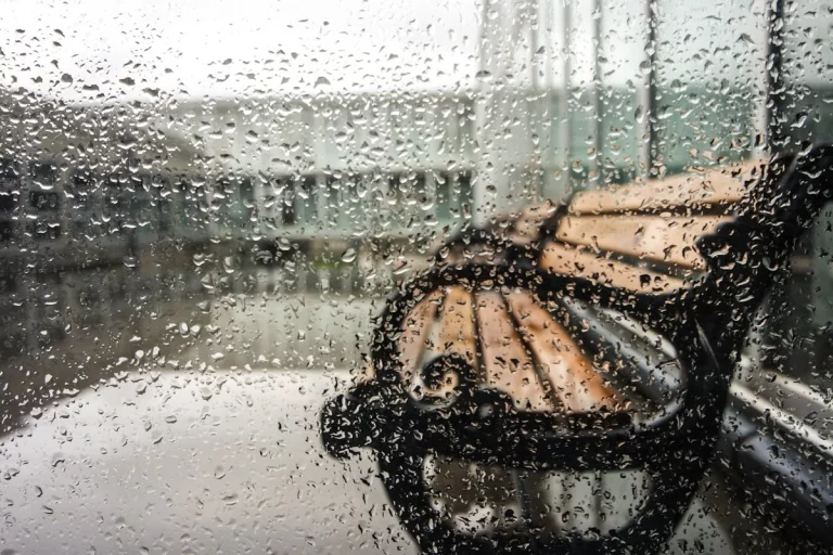 Gotas de chuva escorrendo em uma janela, com foco na superfície molhada, e um banco de praça borrado ao fundo, evocando a sensação da chuva em Florianópolis.