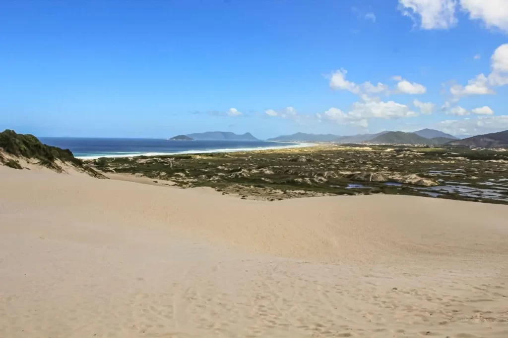 Panorama das dunas de areia da Lagoa da Conceição com vegetação rasteira, lagoa ao longe e morros ao fundo sob céu azul.