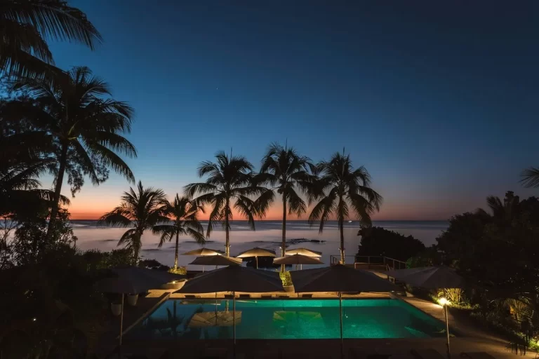Piscina de um hotel ou resort luxuoso em Jurerê Internacional ao anoitecer, com palmeiras silhuetadas contra um pôr do sol vibrante sobre o oceano e espreguiçadeiras iluminadas.