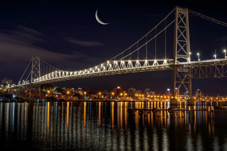 A Ponte Hercílio Luz em Florianópolis iluminada à noite, com uma lua crescente visível no céu escuro, refletindo suas luzes douradas na água e a cidade ao fundo.