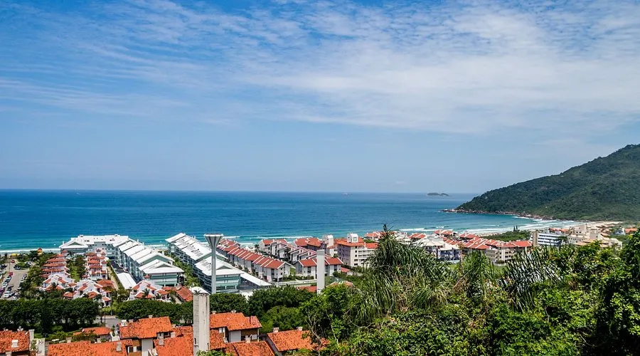 Vista panorâmica da Praia Brava em Florianópolis, SC, com mar, construções e montanhas sob céu azul.