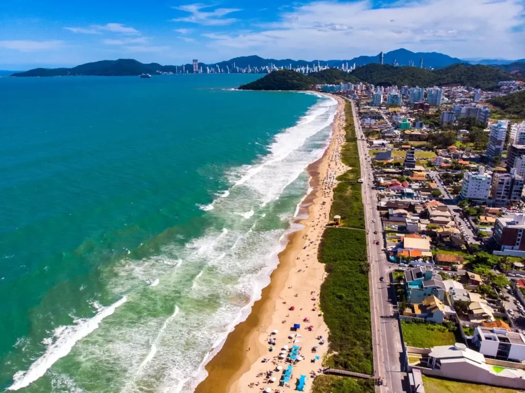 Vista aérea da Praia Brava em Florianópolis, SC, com orla, mar azul intenso, montanhas e arranha-céus ao fundo.
