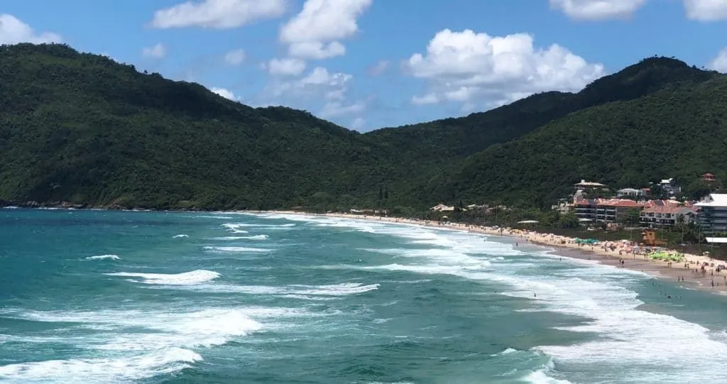 Vista panorâmica da Praia Brava em Florianópolis, SC, com mar agitado, faixa de areia, pessoas e montanhas verdes ao redor.