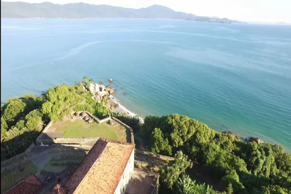 Vista aérea da Praia Forte em Florianópolis, mostrando parte da fortaleza, a faixa de areia, o mar azul e as montanhas ao fundo. Praia histórica em Florianópolis.